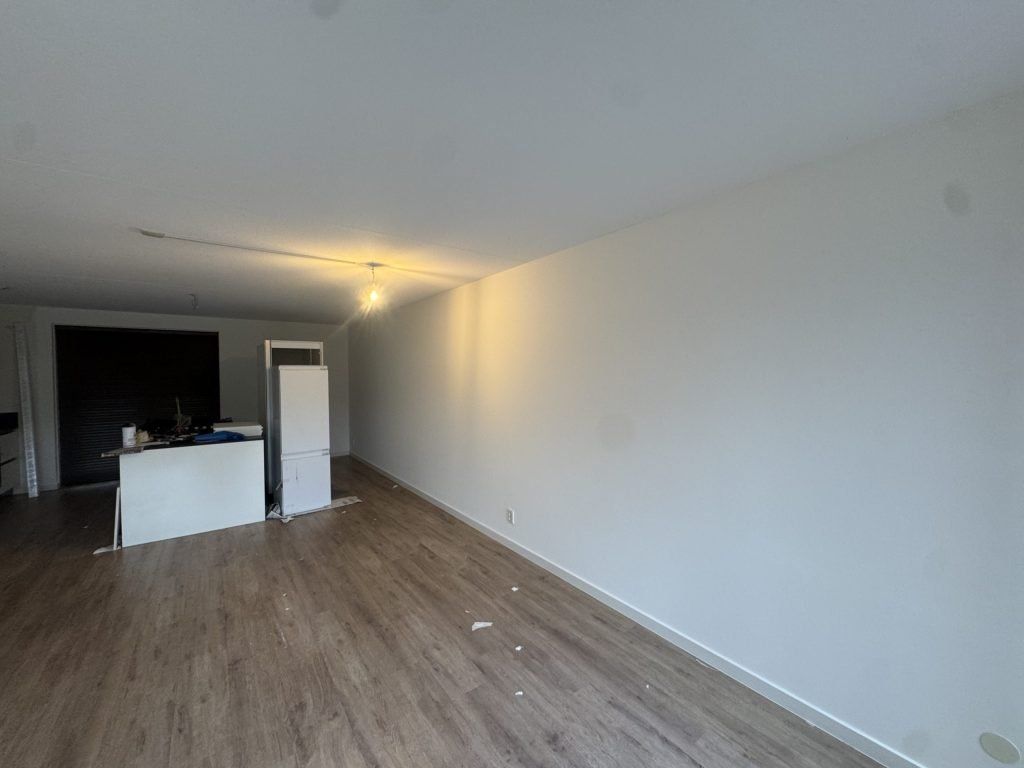 Woonkamer met een houten vloer, een lange witte wand en een keukenblok op de achtergrond