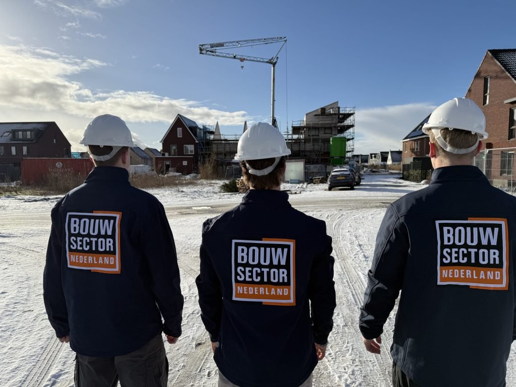 Drie vakmensen van Bouwsector Nederland met witte helm kijken uit over besneeuwde nieuwbouwhuizen met een bouwkraan